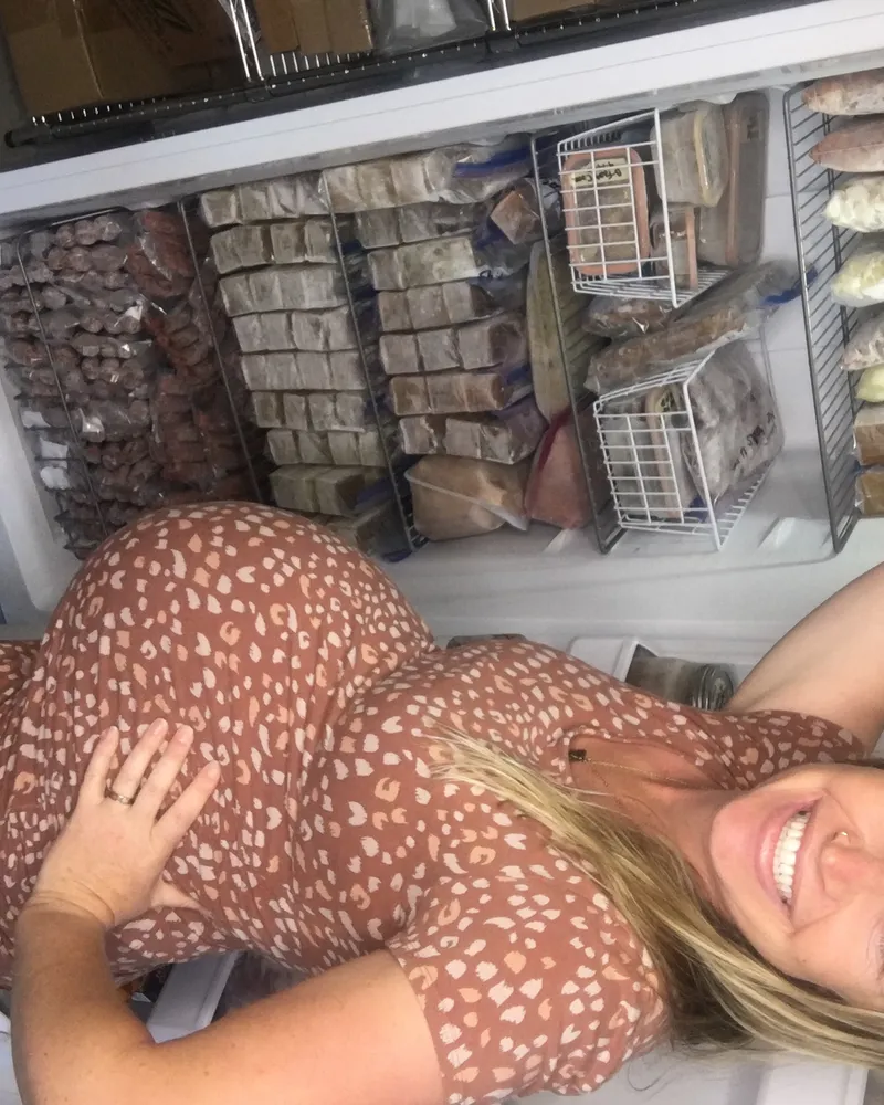 Clare Ramirez pregnant in front of her fully stocked upright freezer packed with home-cooked postpartum meals
