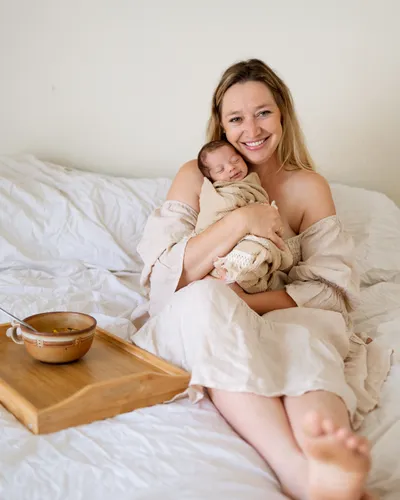 Clare Ramirez resting comfortably in bed with swaddled newborn and warming homemade soup on bedside tray